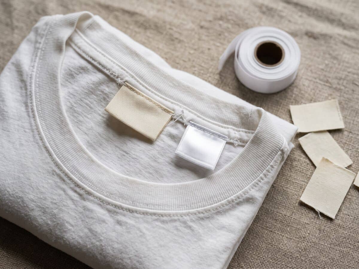 Die Innenseite des Halsausschnitts eines weißen Baumwoll-T-Shirts mit einem leeren gewebten Nackenetikett und einem in die Naht eingenähten Pflegeetikett aus Satin, daneben eine Rolle Etikettenband.