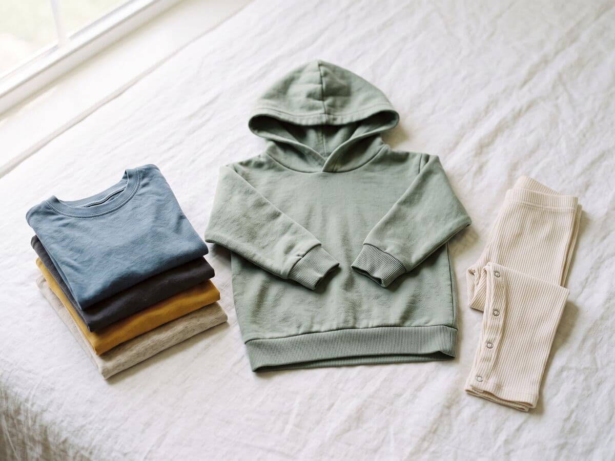 A sage children's hooded sweatshirt with no drawstrings in the hood, laid flat beside folded toddler t-shirts and snap-fastened leggings.