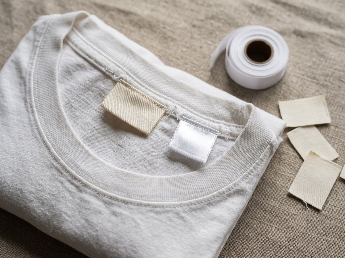 The inside neck of a white cotton t-shirt with a blank woven neck label and a satin care label stitched into the seam, beside a roll of label tape.