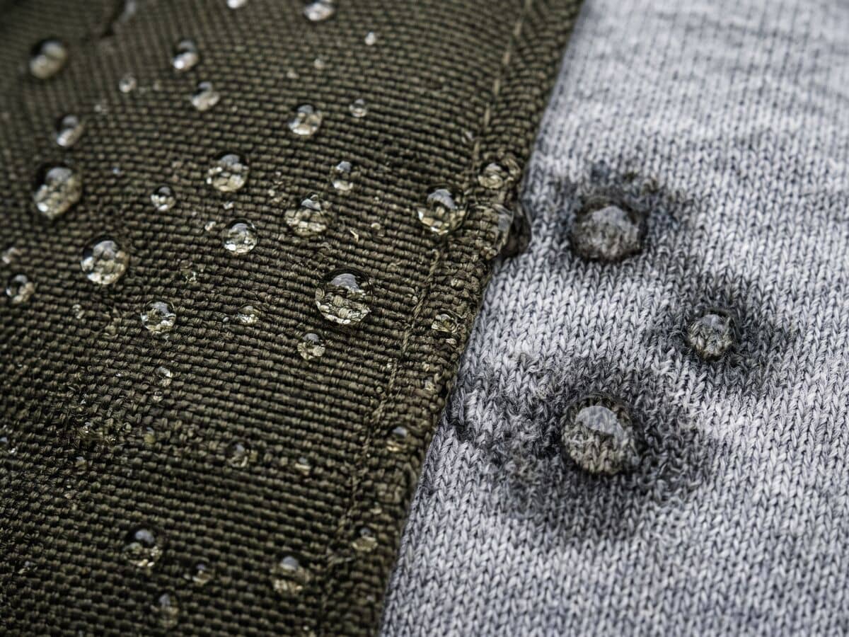 Gotas de agua que forman perlas sobre el tejido hidrófugo de una chaqueta verde oliva, mientras otras gotas empapan un punto de algodón gris a su lado.