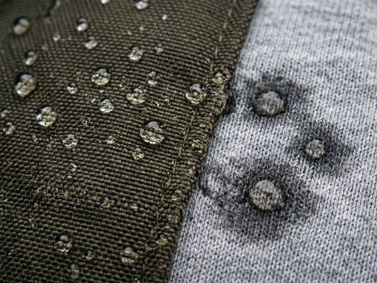 Gotas de agua que forman perlas sobre el tejido hidrófugo de una chaqueta verde oliva, mientras otras gotas penetran en un tejido de punto de algodón gris a su lado.