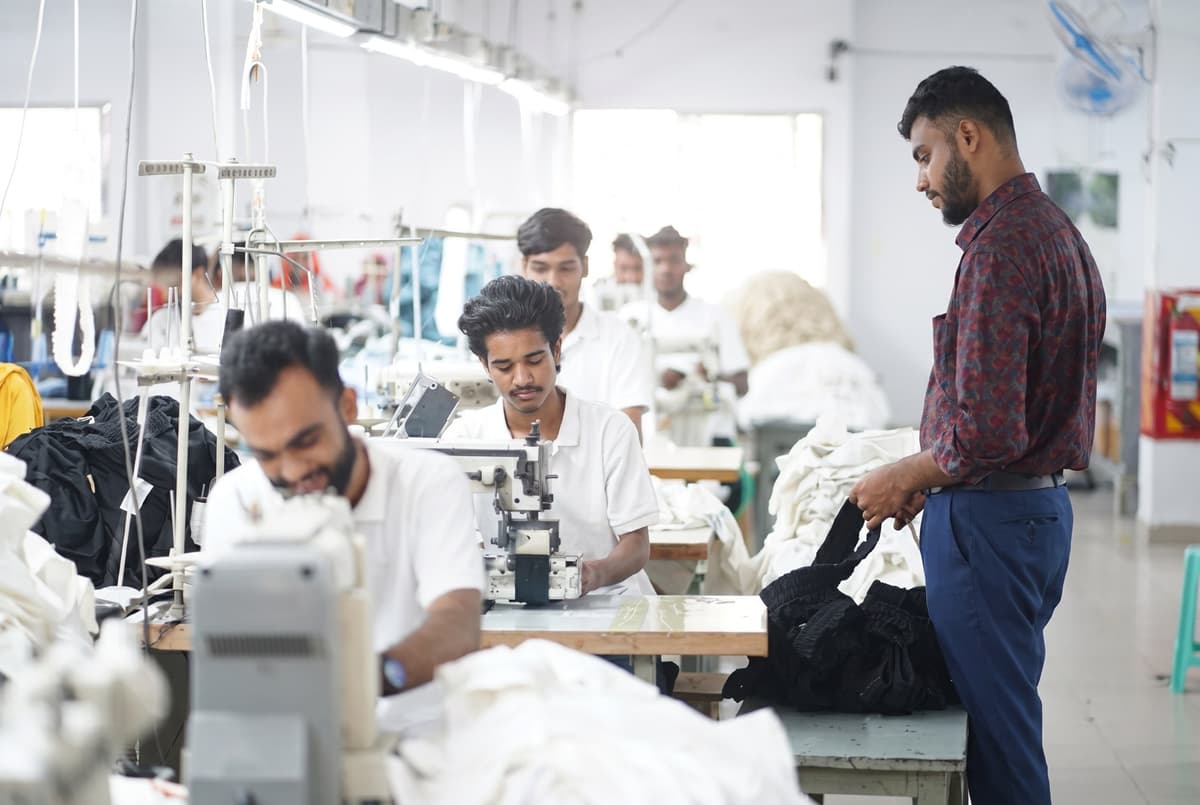 Workers at sewing stations on the Collective Studio line, a colleague in a checked shirt reviewing the stitching.